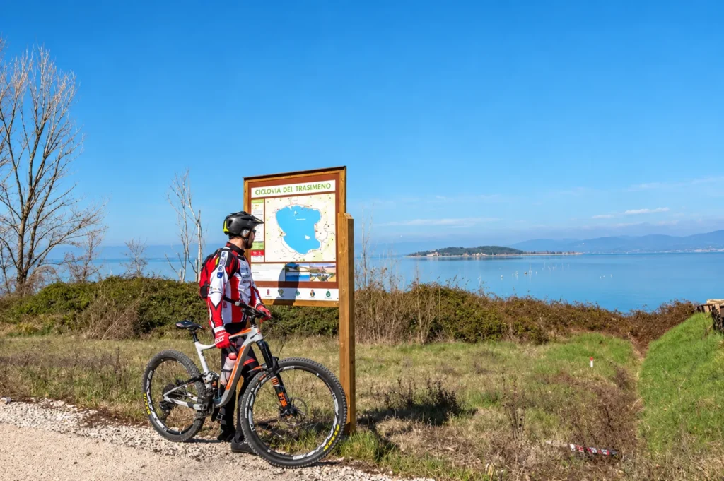 Ciclista con mountain bike accanto al pannello della Ciclovia del Trasimeno, con il lago sullo sfondo