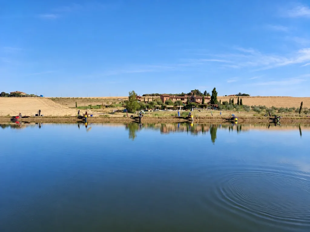 See der Fishing Accademy in Paciano mit Anglern am Ufer und Bauernhäusern dahinter