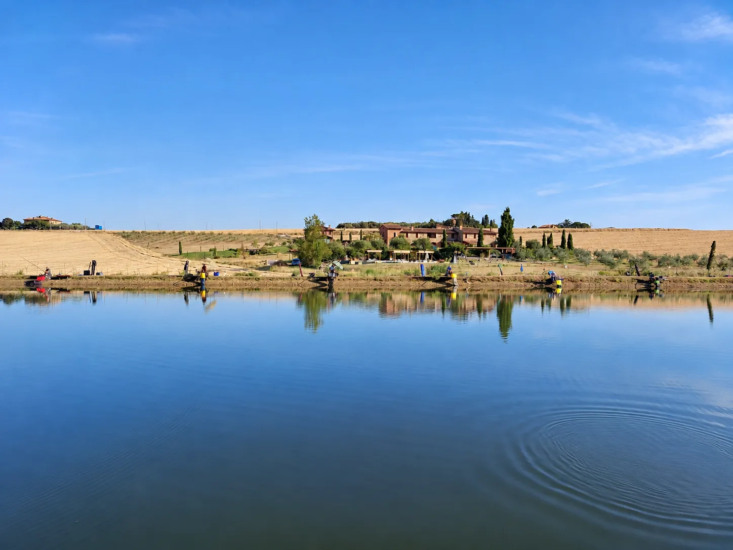 See der Fishing Accademy in Paciano mit Anglern am Ufer und Bauernhäusern dahinter