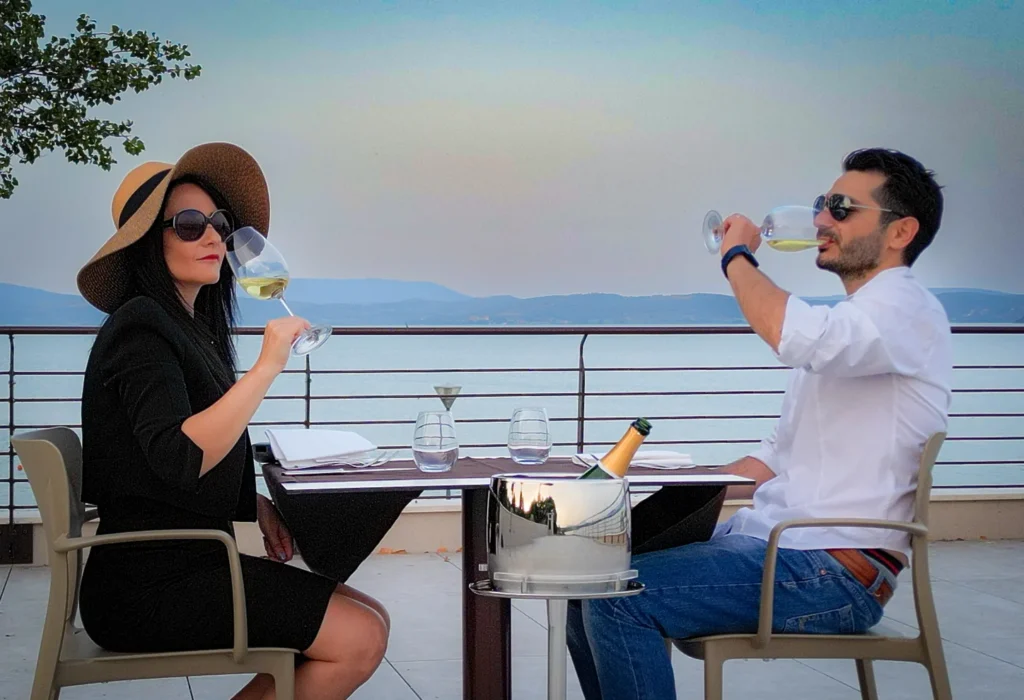 Deux personnes dégustent du vin blanc sur une terrasse face au lac, avec une bouteille dans un seau à glace à côté de la table.