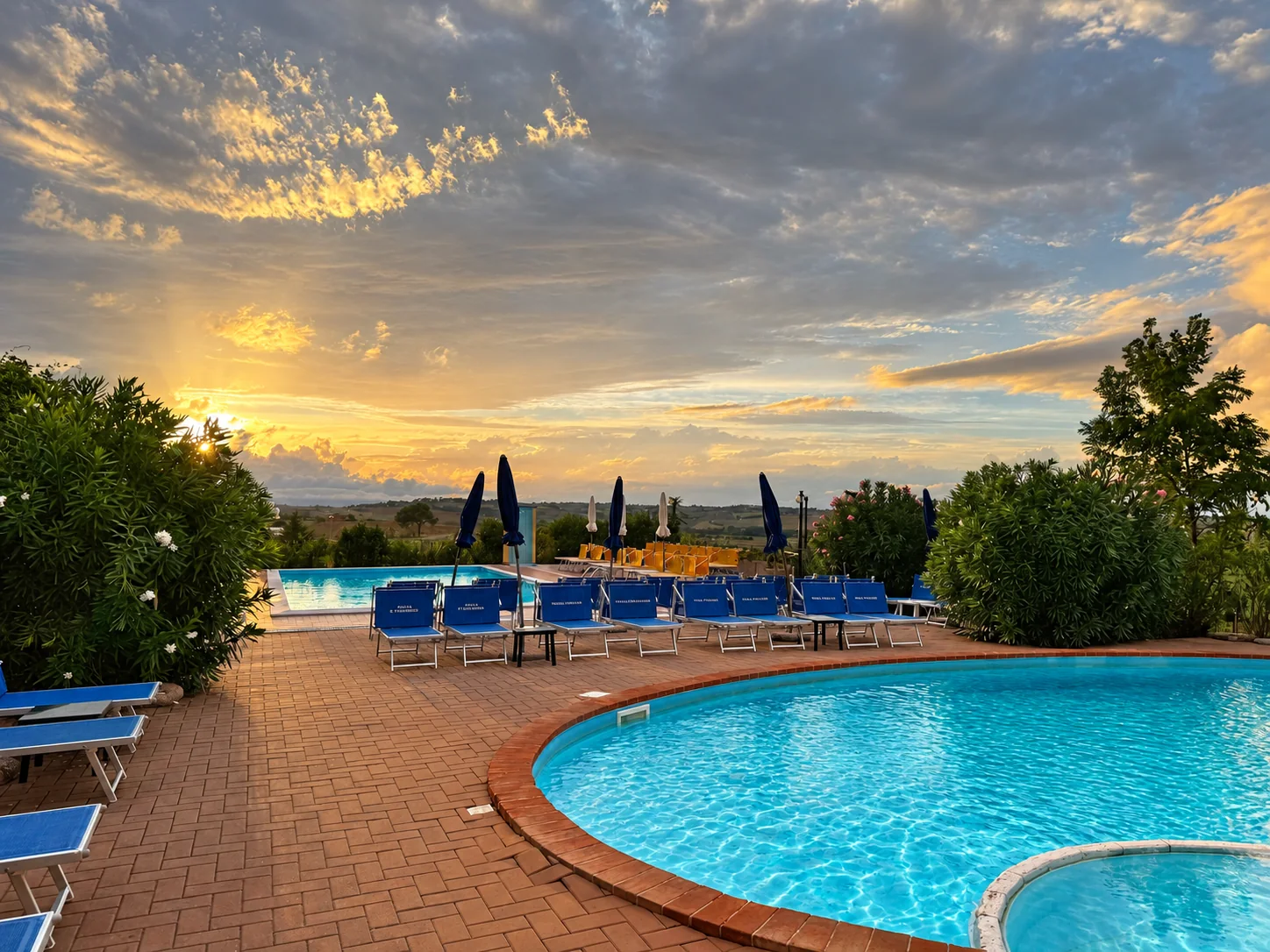 Le due piscine di Tenuta Badia con solarium, oleandri e cielo dorato al tramonto sulla campagna umbra.