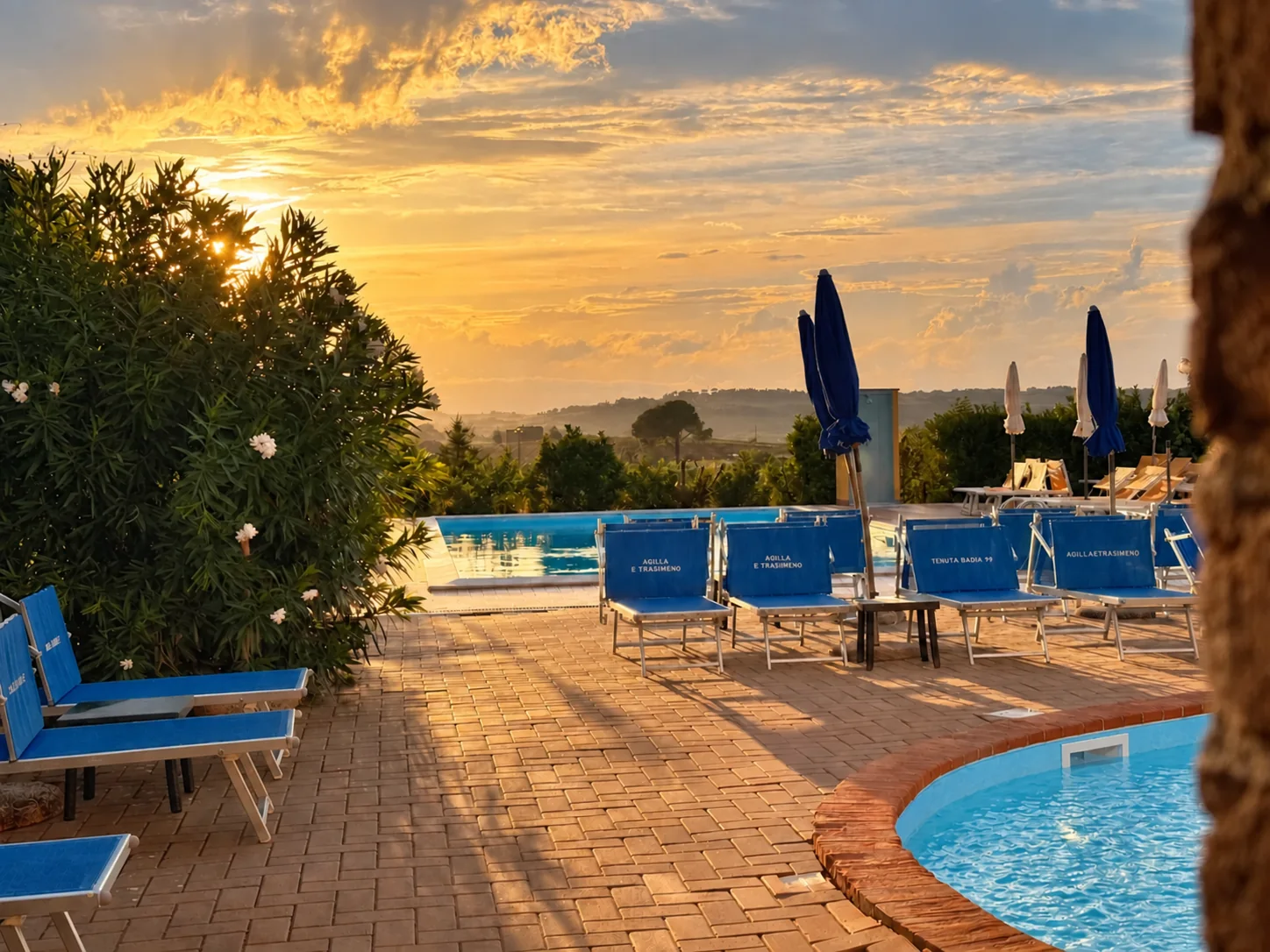 Lettini blu e ombrelloni tra le piscine di Tenuta Badia, illuminati dalla luce dorata del tramonto.
